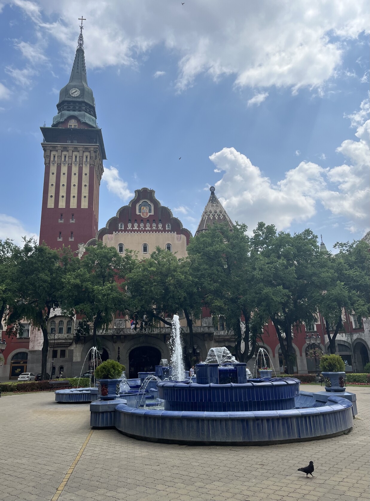 Синий комплекс фонтанов, за которым городская ратуша, прячущаяся за листвой деревьев 
